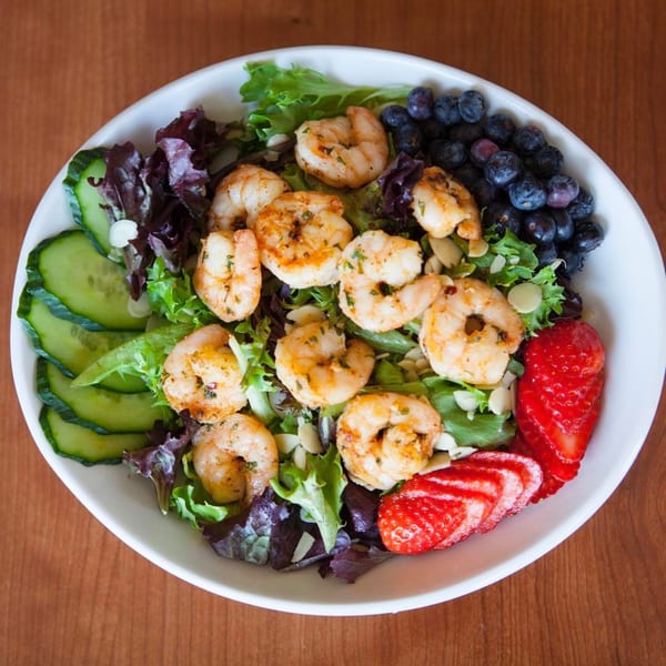 A vibrant shrimp salad with mixed greens, blueberries, strawberries, cucumbers, and almonds in a white bowl.