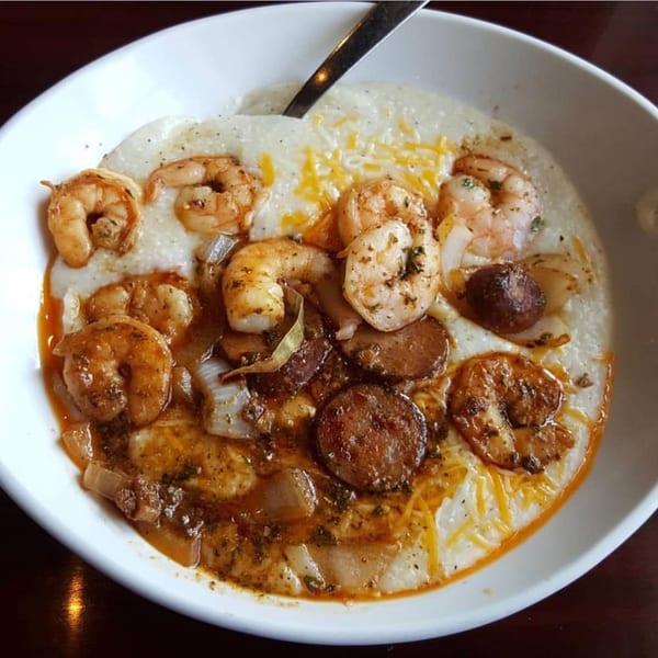 A white bowl of creamy grits topped with shrimp, sausage, onions, and melted cheese.