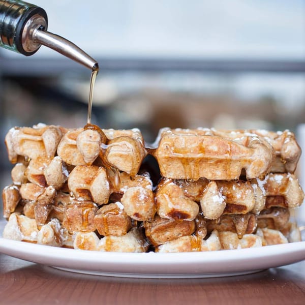 Syrup pouring from a metal dispenser onto two stacks of golden-brown waffles dusted with powdered sugar.